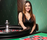 Una crupier sentada ante una mesa de ruleta que mira a la cámara, un servicio que Casino777 aun no ofrece.